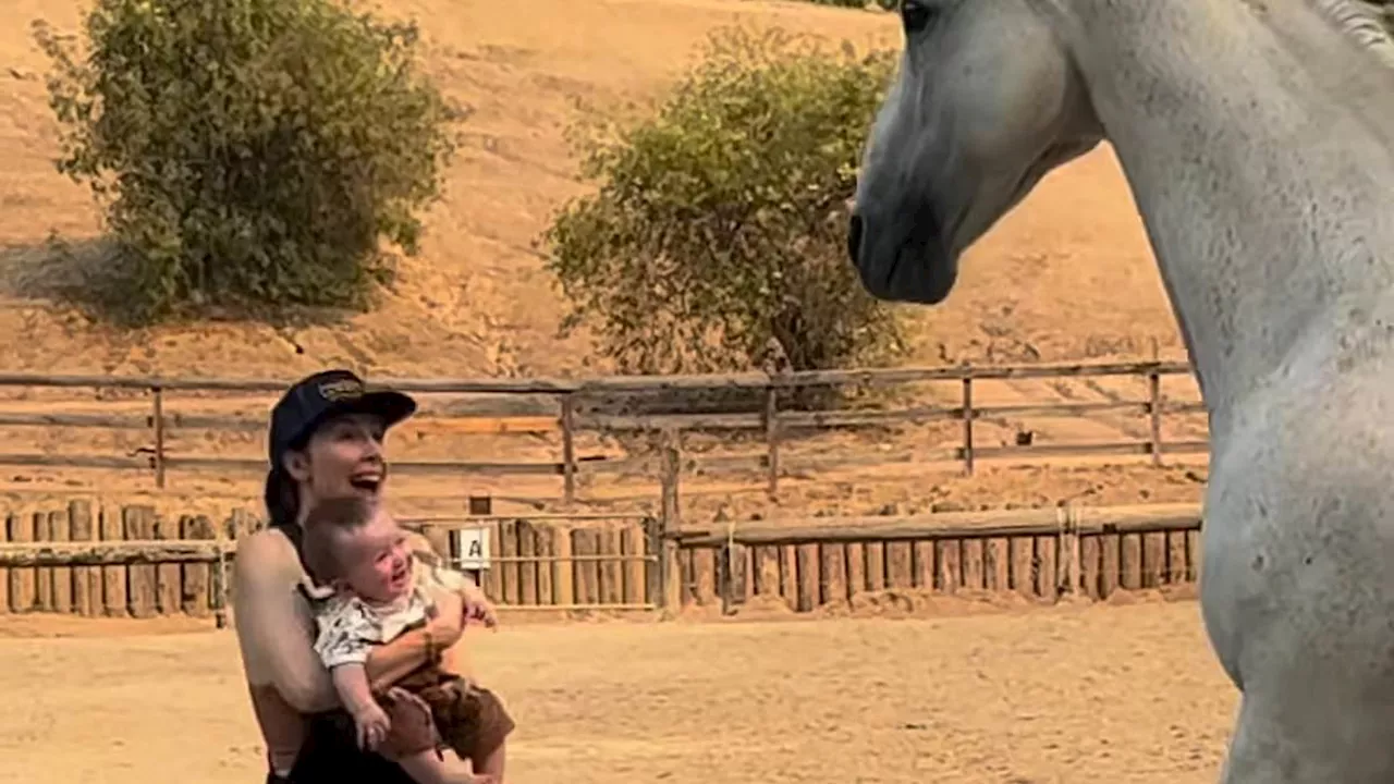 Whitney Cummings holds her baby up in front of a bucking horse